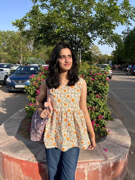 Woman wearing a sleeveless cotton top with marigold floral print, standing outdoors near flowers, styled with blue jeans and a printed tote bag.