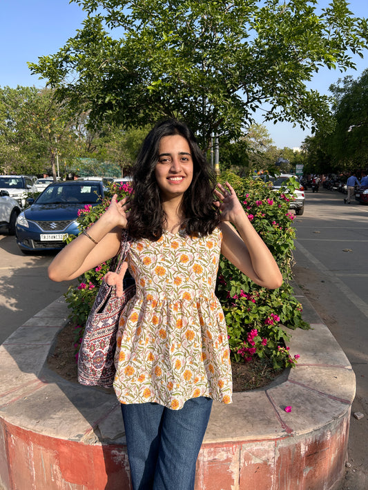 Woman wearing a sleeveless cotton top with marigold floral print, standing outdoors near flowers, styled with blue jeans and a printed tote bag.