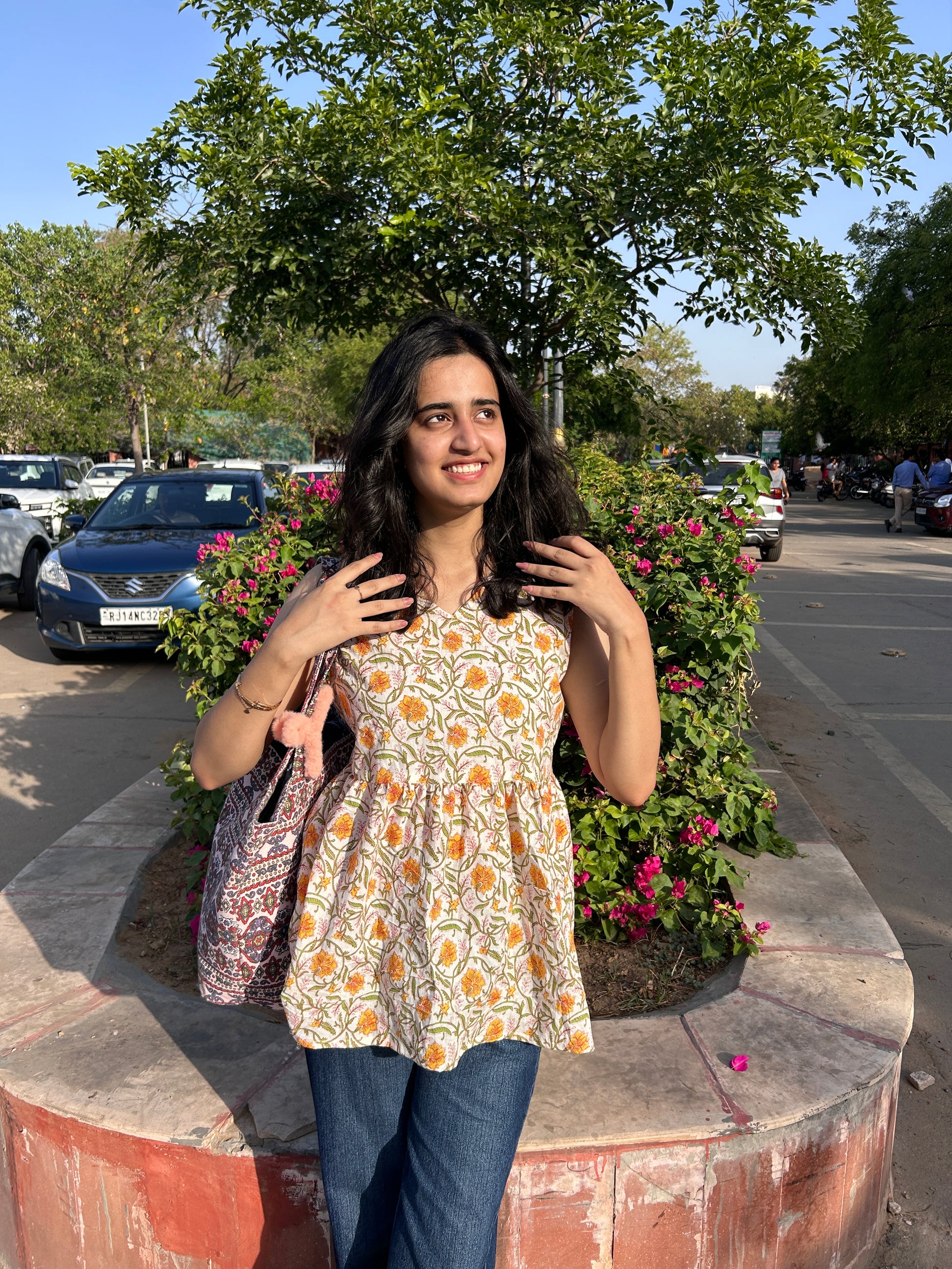 Woman wearing a sleeveless cotton top with marigold floral print, standing outdoors near flowers, styled with blue jeans and a printed tote bag.