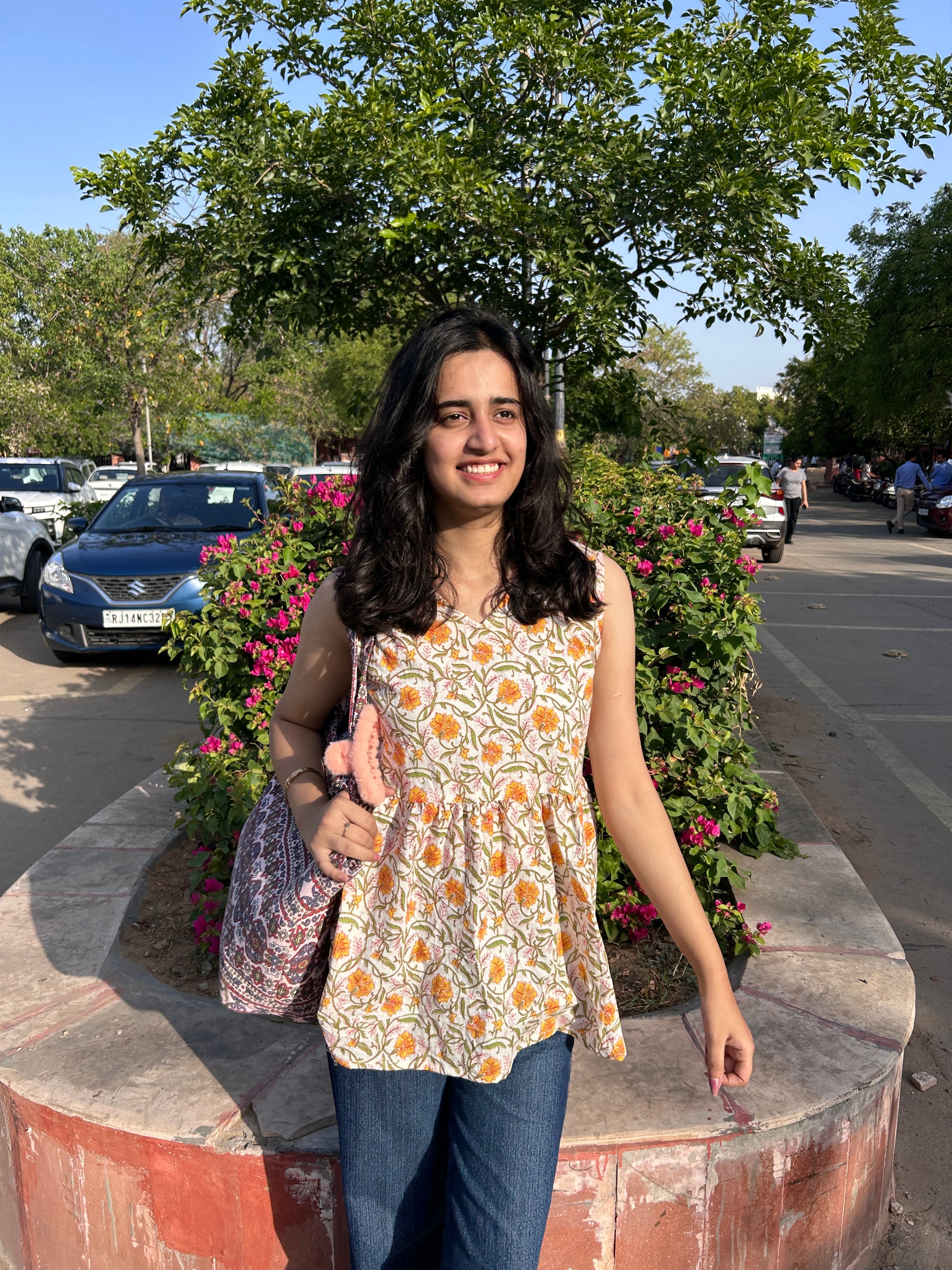 Woman wearing a sleeveless cotton top with marigold floral print, standing outdoors near flowers, styled with blue jeans and a printed tote bag.